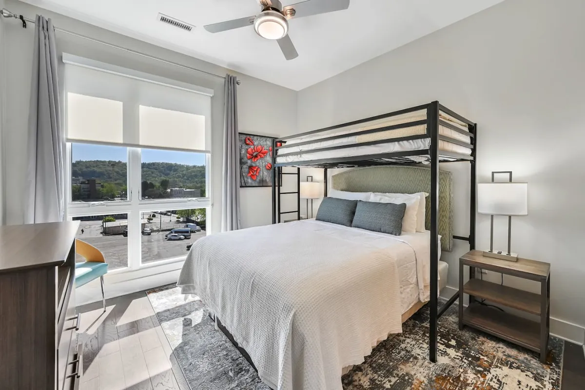 A cozy, modern bedroom featuring a lofted bed bathed in natural light from a large window, decorative wall art, and a nightstand with a lamp. The space exudes both comfort and privacy, ideal for restful nights or quiet reading.