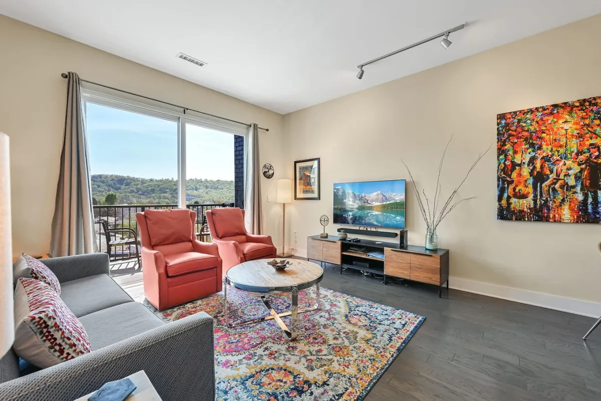 A bright and cozy living room featuring plush red armchairs, a colorful area rug, wall-mounted artwork, and a television. The space is arranged for both relaxation and entertaining, with a glimpse of the balcony in the background, hinting at access to outdoor views and natural light.