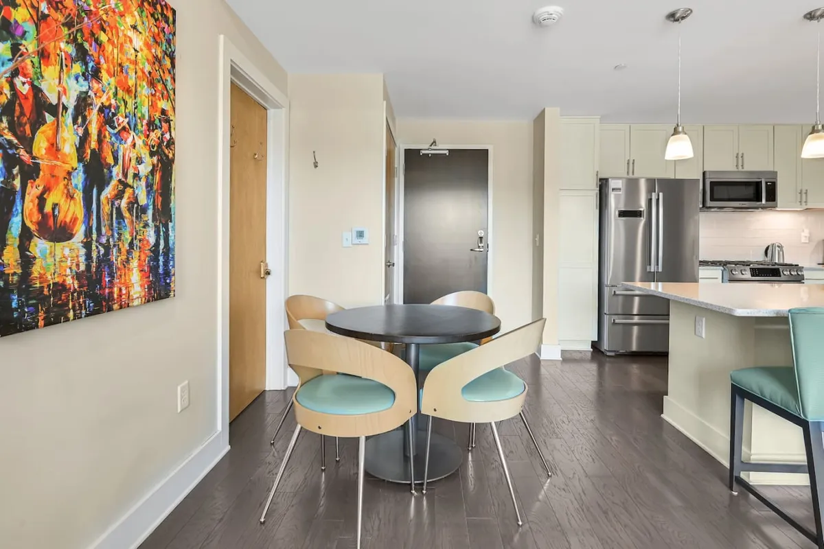 A cozy dining area with a round table set beneath colorful artwork, seamlessly connected to a view of the modern kitchen and appliances. The space feels artistic and inviting, perfect for intimate meals or cheerful gatherings.