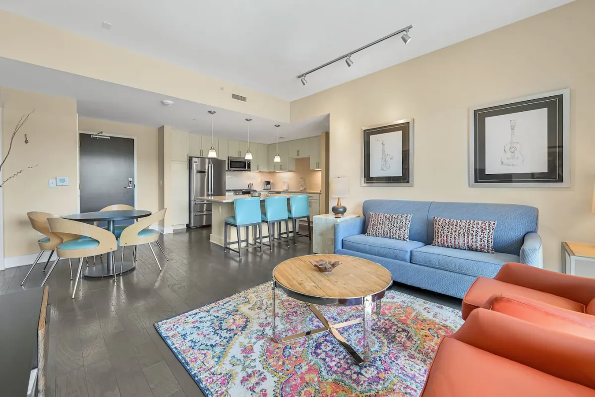 The image features a stylish open-concept living space with a plush blue sofa, a vibrant and colorful rug, a modern dining area with chairs, and a sleek kitchen in the background. The scene is bright and inviting, perfect for relaxing, socializing, or entertaining guests, all set within a premium downtown Asheville condo.