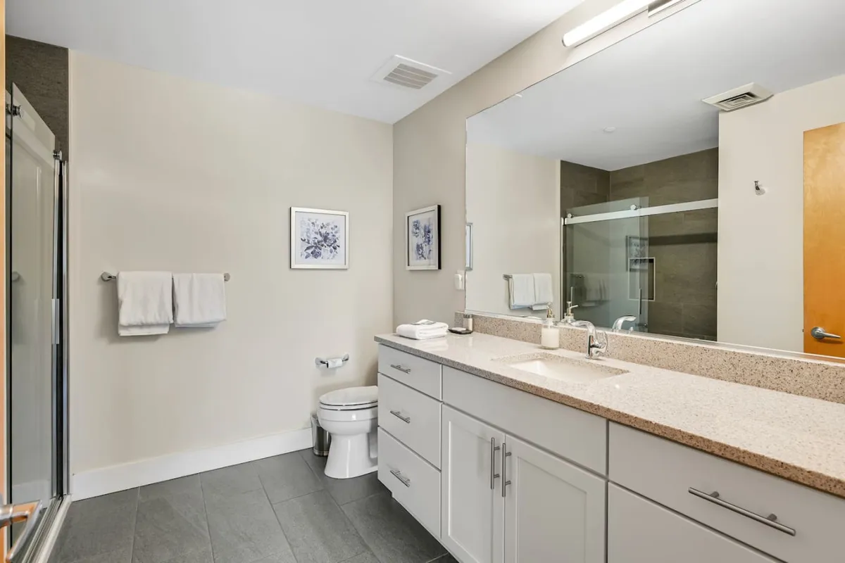 A spa-inspired bathroom featuring a double sink vanity, expansive mirror, glass shower enclosure, and tasteful wall art. The calming ambiance and practical layout make it ideal for rejuvenation and self-care rituals.
