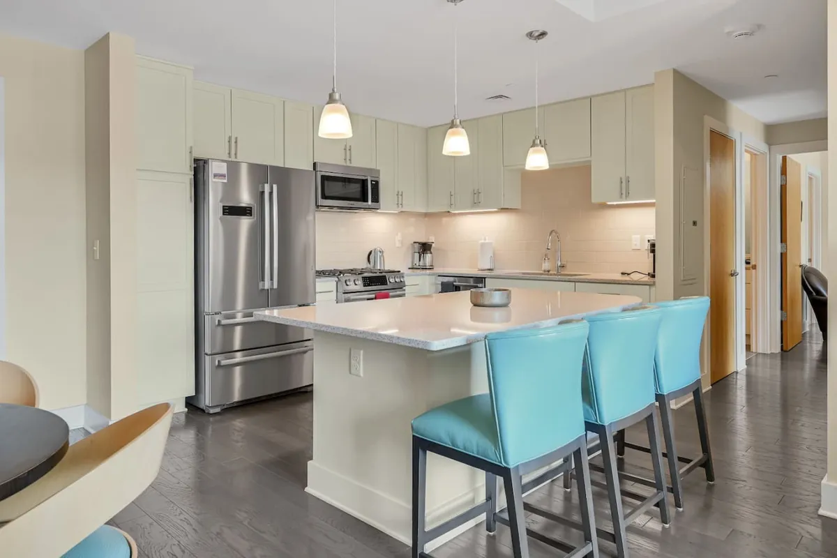This image features a contemporary kitchen with a spacious island countertop, stainless steel appliances, elegant pendant lighting, and inviting blue bar stools. The overall ambiance is welcoming and modern, with thoughtful details that highlight functionality and style.