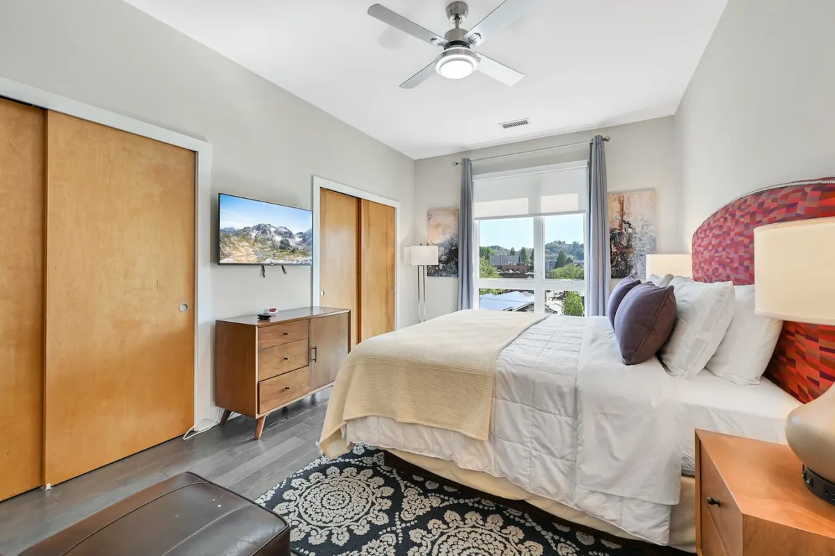A cozy bedroom scene featuring a comfortable bed, a television, a nightstand, and a window that frames a city view. The room feels restful and modern, with artwork adding a decorative touch.
