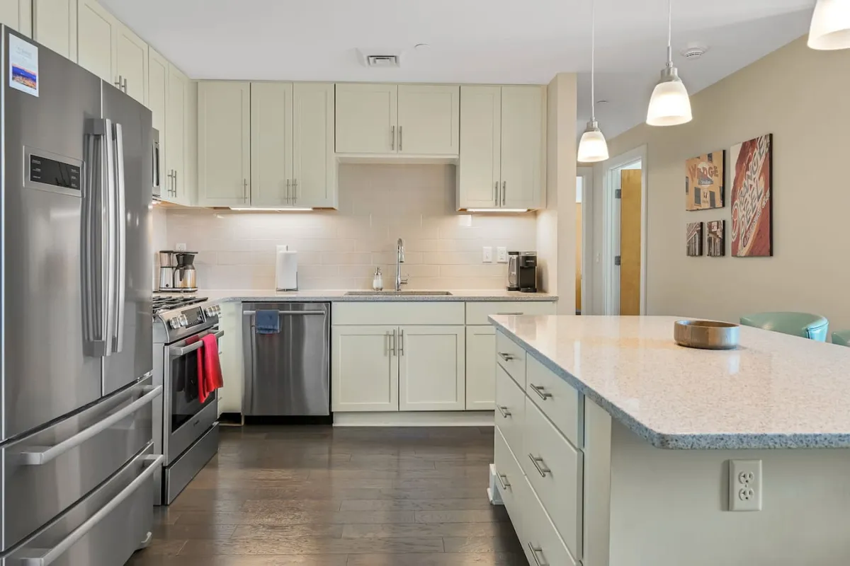 A brightly lit kitchen centered around a granite-topped island, featuring a stainless steel refrigerator, ample workspace, and creative wall art. The layout is open and conducive to both cooking and entertaining, while the mix of textures adds visual interest.