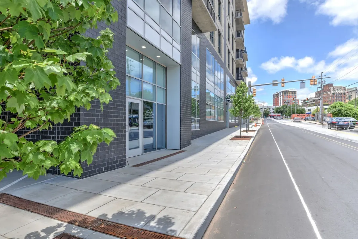 This image shows the modern exterior of the condo building, featuring large glass windows, well-maintained landscaping with trees, and a bustling sidewalk. The setting is vibrant and urban, suggesting proximity to cafes, shops, and community spaces.