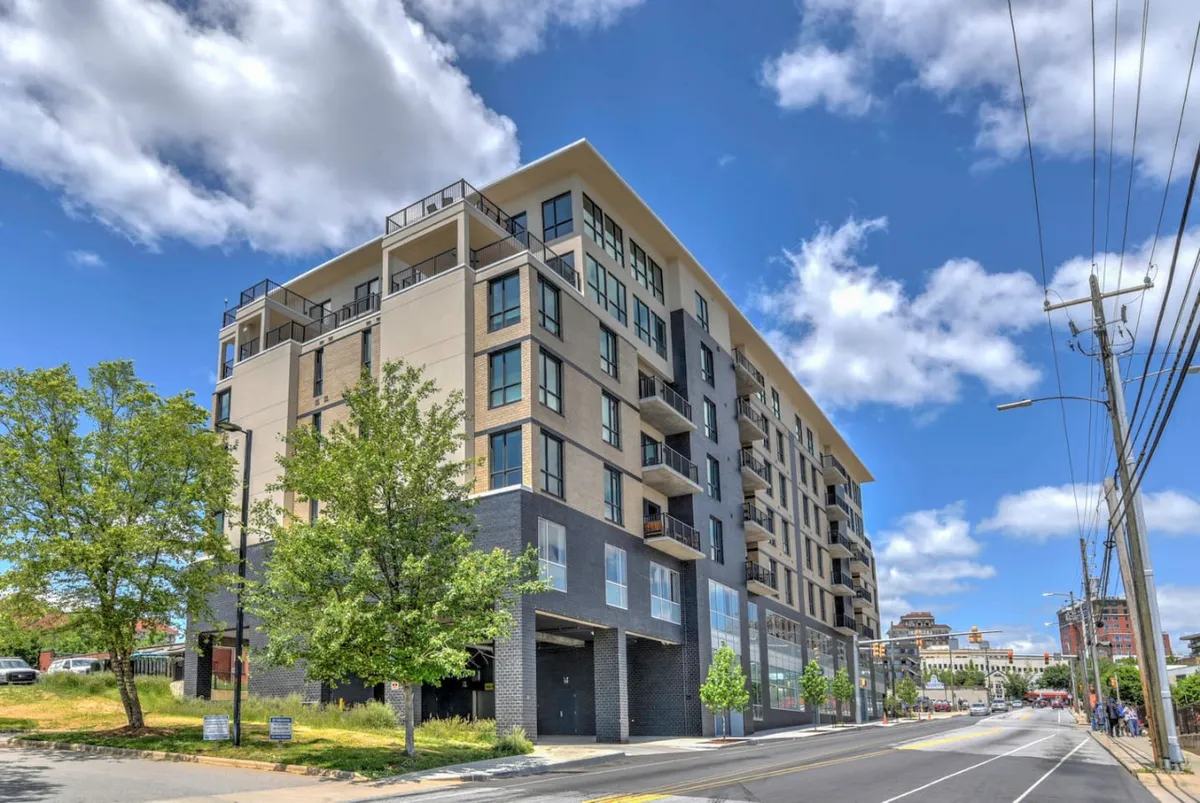 This image features the sleek, modern exterior of a condominium building, highlighted by expansive windows, multiple balconies, and lush green landscaping. The urban surroundings and architecture signal a vibrant city environment, ready to be explored.