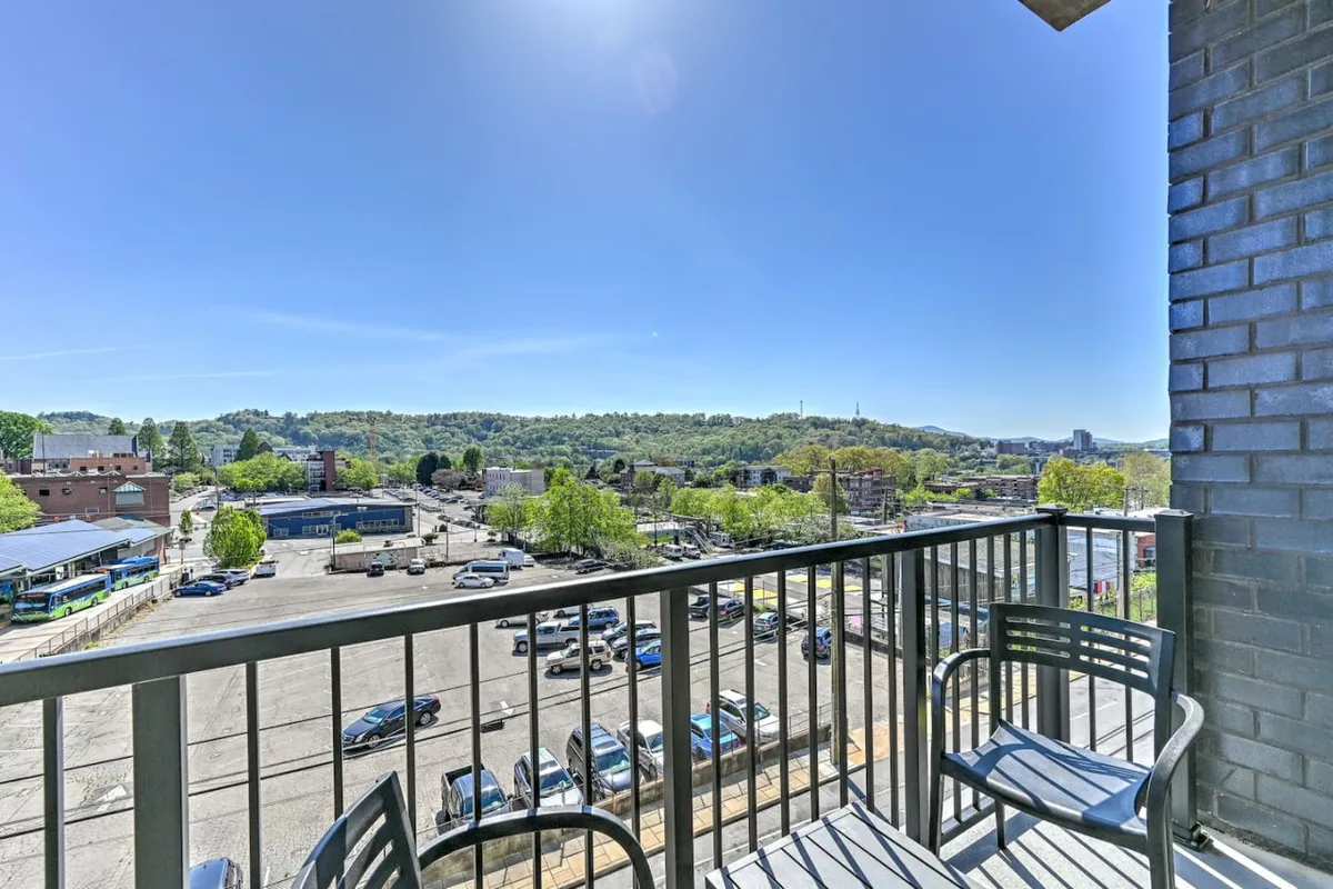 From the balcony, you see a cityscape with parked cars, a mountain backdrop, and the skyline stretching beyond. The view includes a glimpse of the dedicated parking area, connecting the image to convenience and security.