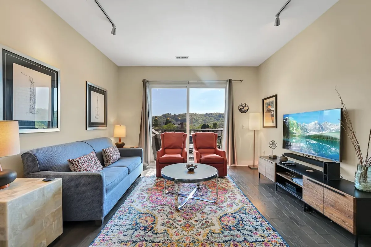 A welcoming living area with a comfortable sofa, red chairs, a central coffee table perfect for games or drinks, a television, and large windows offering city views. The open layout seamlessly blends social and relaxation spaces.