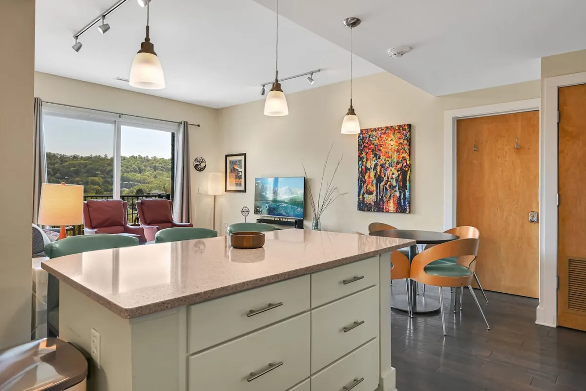 This image showcases a bright, open kitchen area with a central island, modern seating, wall art, and expansive windows that offer views to the outside. The scene suggests an inviting spot for casual meetings, a quick meal before heading out, or unwinding with a city view.